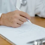 Profissional de saúde com jaleco branco sentado em mesa de escritório, segurando documento ou prontuário, simbolizando atenção à saúde e atendimento médico.