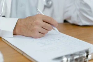 Profissional de saúde com jaleco branco sentado em mesa de escritório, segurando documento ou prontuário, simbolizando atenção à saúde e atendimento médico.